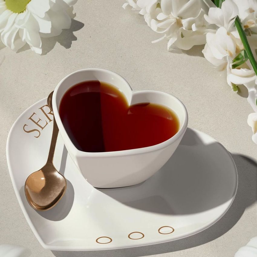 Heart-shaped cup mockup filled with dark liquid on a matching plate with a gold spoon, surrounded by white flowers.