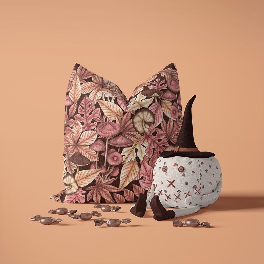 Halloween pillow mockup with a fall leaves pattern next to a plush toy pumpkin wearing a witch hat, candies scattered in front.