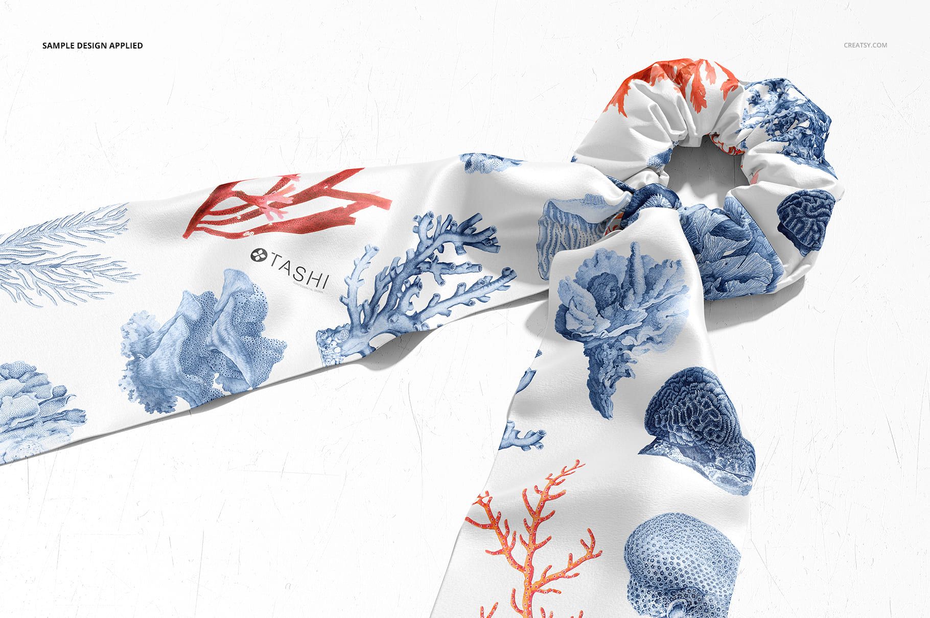 Fashion accessory mockup displaying a scrunchie scarf with ocean-inspired blue and red coral designs on white.