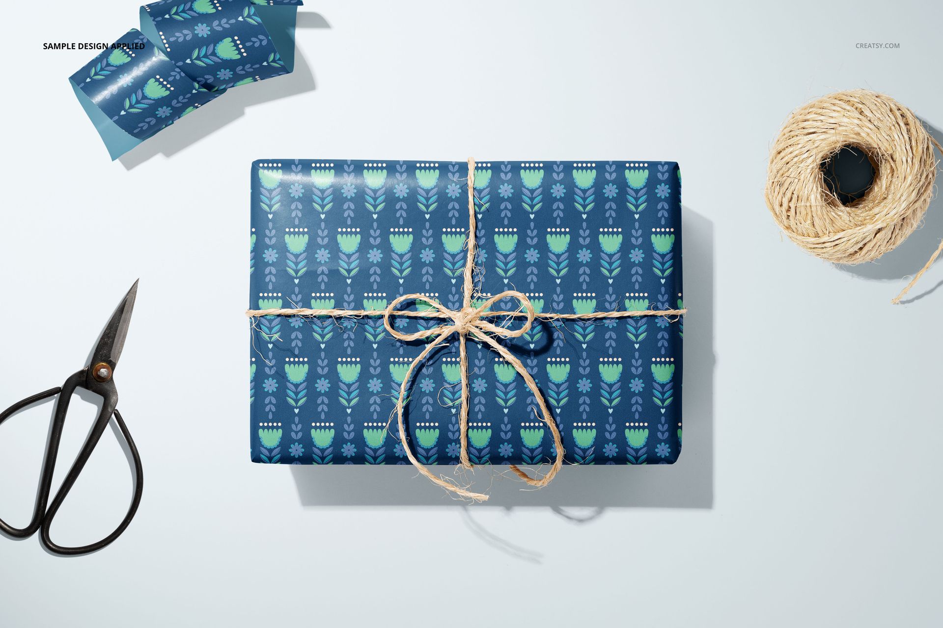Gift wrapping paper mockup featuring a blue geometric pattern, twine bow, scissors, and spool of string on a white surface.