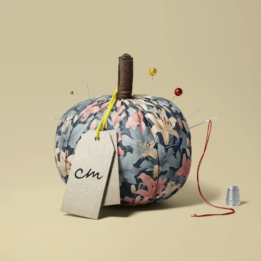 Fabric pumpkin mockup with a floral pattern, pins, and a blank tag on a beige background with a thimble and thread.
