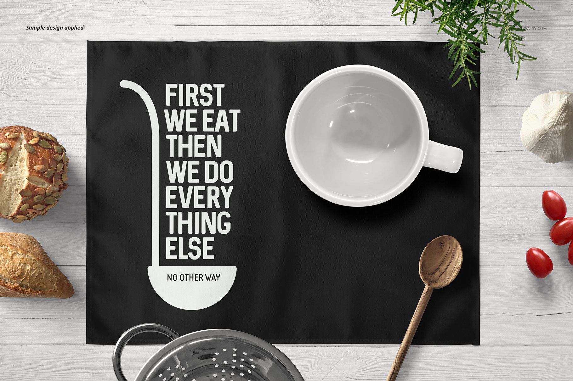 Rectangular black placemat mockup with bold white text, displayed on a light wooden table with bread, tomatoes, and herbs.