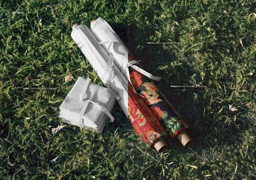 Fabric rolls and folded stacks with grid and floral patterns arranged on green grass, viewed from above.