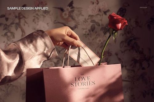 PSD mockup showing a hand adding a card to a pink gift bag decorated with a red rose, in front of floral wallpaper.