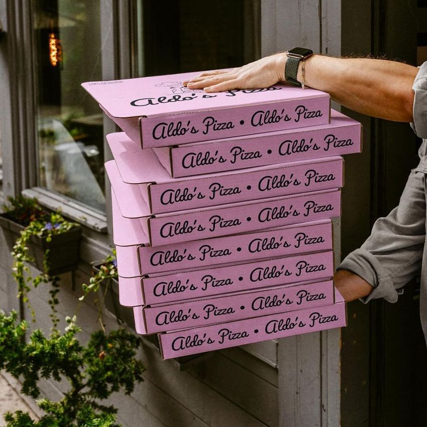 Person carrying multiple pink pizza box mockups with cursive text, standing near a window and plants in an outdoor setting.