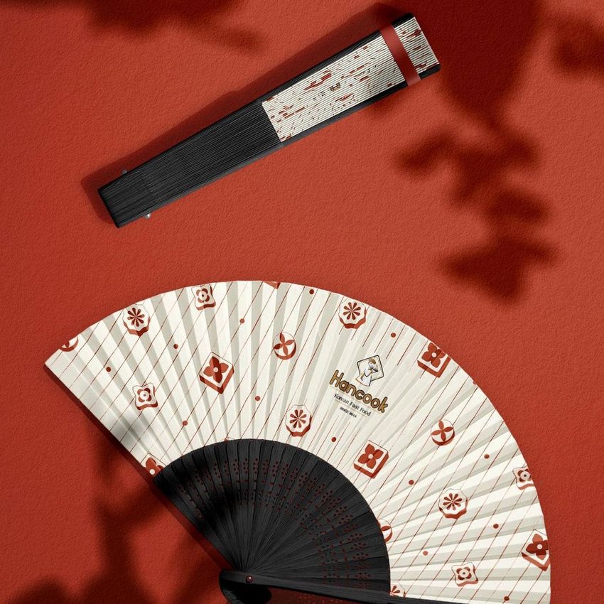 Fabric hand fan mockup with a black handle and patterned white fabric, displayed on a red textured background with geometric motifs.