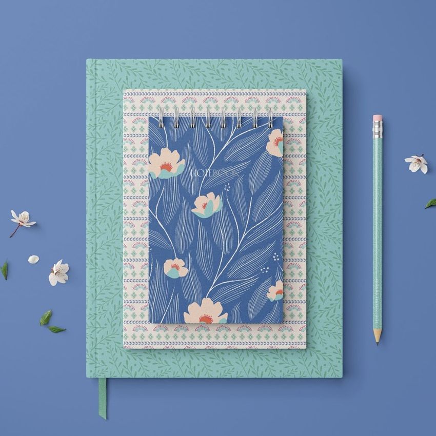 Stationery set mockup featuring a blue floral notebook, patterned paper, and a green journal on a blue background with a pencil.