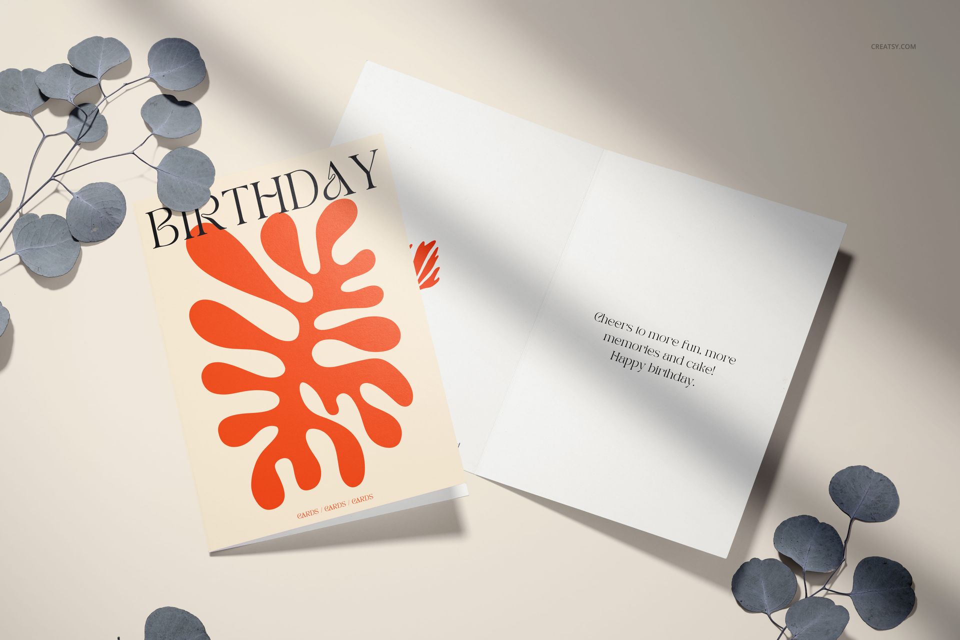 5x7 greeting card PSD mockup showing cover with bold orange pattern and open interior, styled with eucalyptus leaves.
