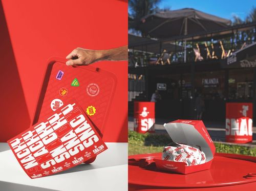 Red fast food tray with bold white lettering and colorful stickers, next to a burger box and wrapper outdoors.