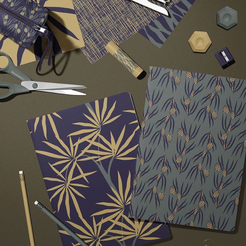 Stationery mockup featuring two classic A4 notebooks with floral and botanical patterns, surrounded by pencils, scissors, and paper props on a dark surface.