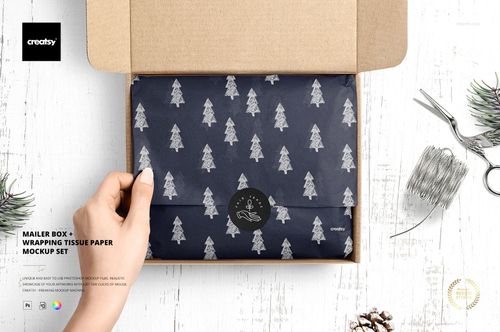 Mailer box PSD mockup displaying folded navy tissue paper with tree motifs, hand holding the lid, and packaging props nearby.