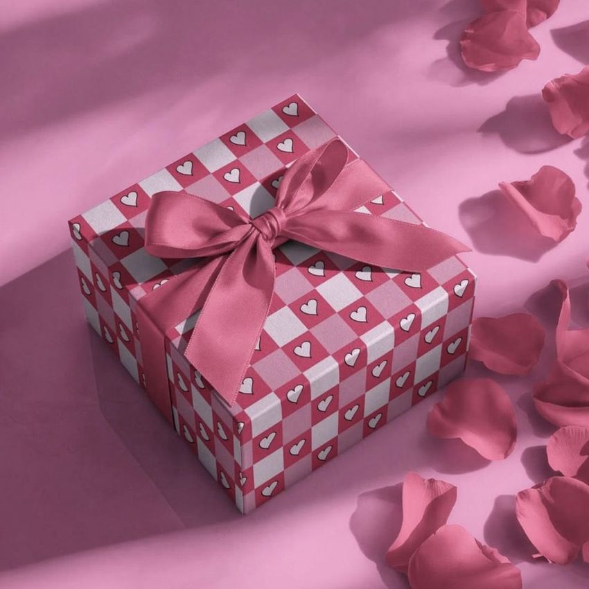 Square gift box mockup with a pink ribbon, heart pattern wrapping, and scattered rose petals on a pink surface.