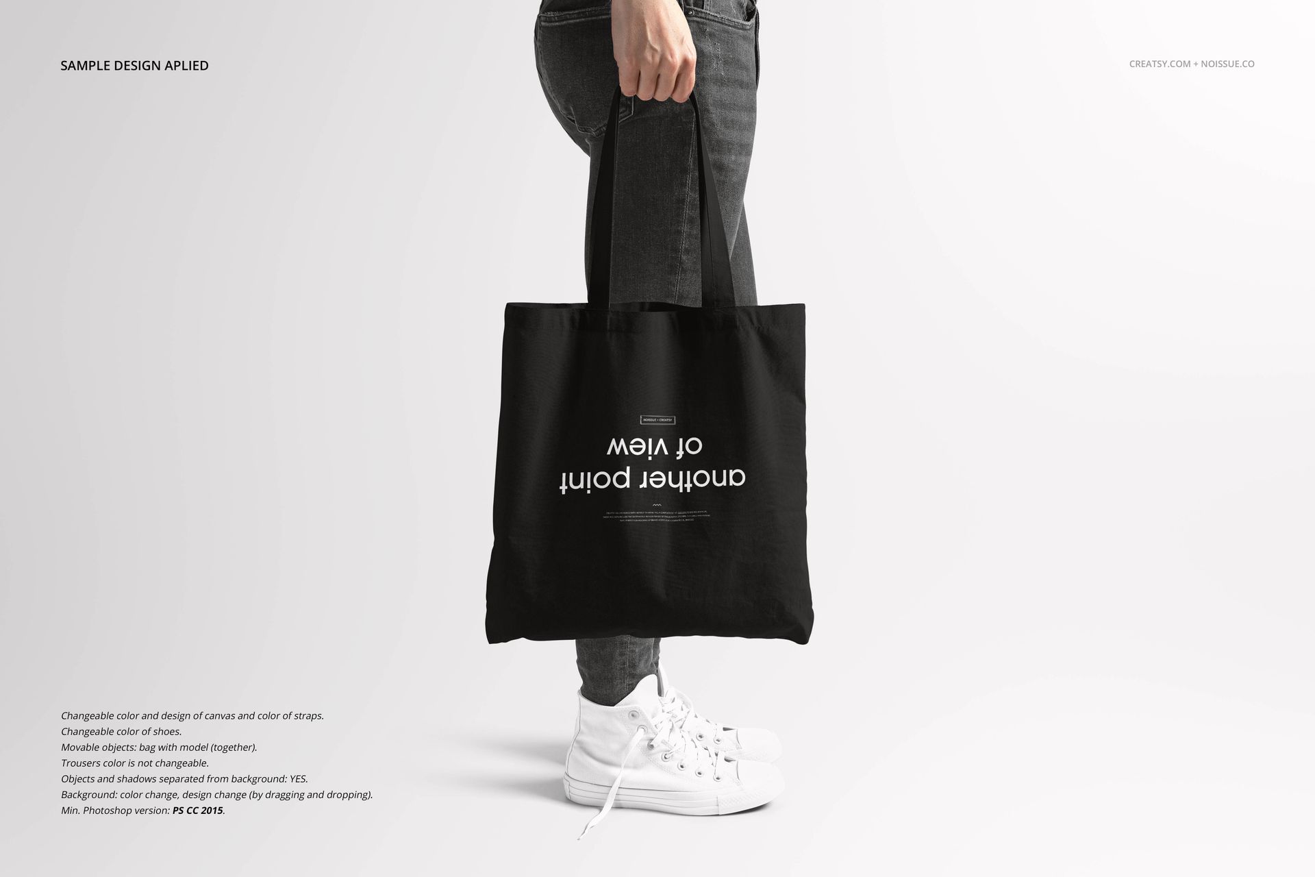 Black tote bag mockup held by a person wearing dark jeans and white sneakers, shown on a minimalist background.