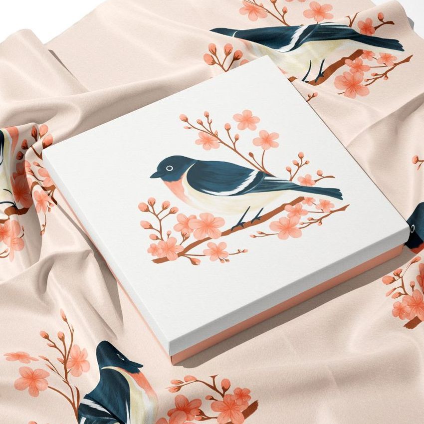 Silk scarf box mockup featuring a square box with a bird and pink floral pattern, placed on matching patterned fabric.