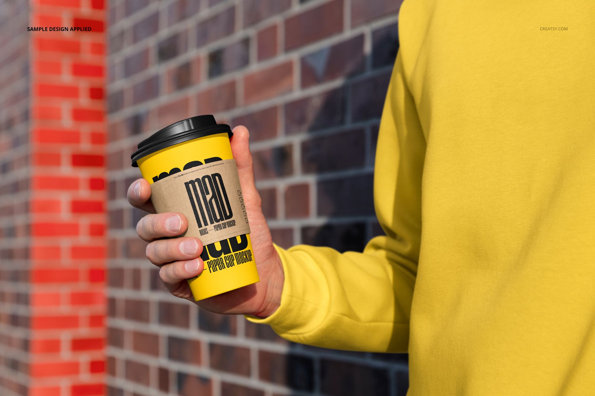 Close-up of a person in a yellow sweatshirt holding a yellow paper cup mockup with black lid and graphic text.