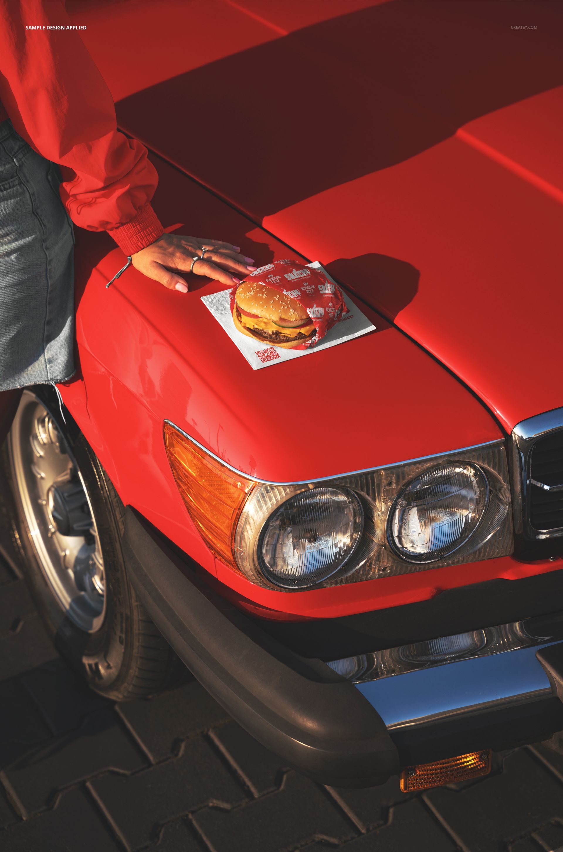 PSD mockup showing a burger and napkin positioned on the front hood of a red classic car, photographed from above.