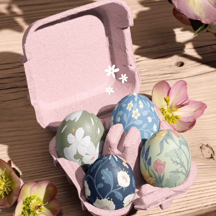Egg carton mockup with four decorated eggs featuring floral patterns, surrounded by hellebore flowers on a wooden surface.