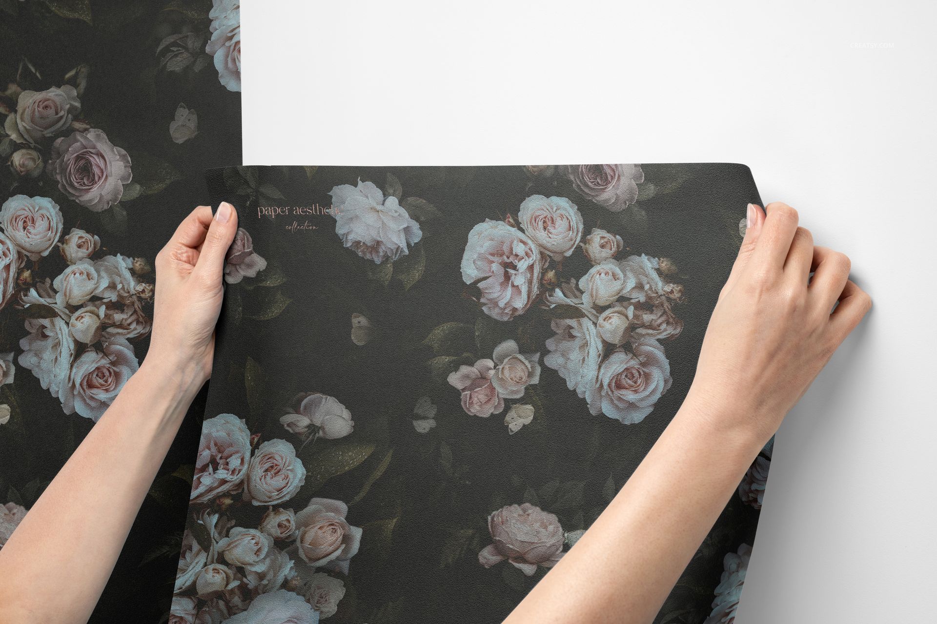Close-up of hands positioning a dark floral non-woven vinyl wallpaper mockup, showing detailed rose designs and texture.