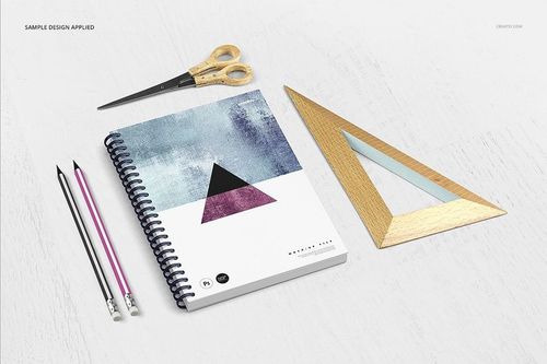 Front view of a notebook mockup featuring abstract cover art, next to gold scissors, pencils, and a wooden triangle ruler.