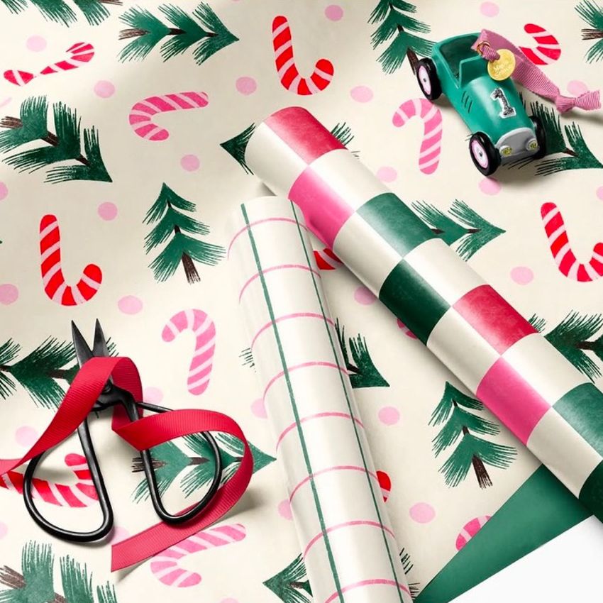 Gift wrapping paper mockup featuring Christmas-themed patterns with candy canes, pine branches, and pink dots, shown with scissors, ribbon, and a toy car.