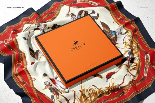Front view of an orange silk scarf box mockup resting on a decorative scarf featuring birds and equestrian elements.