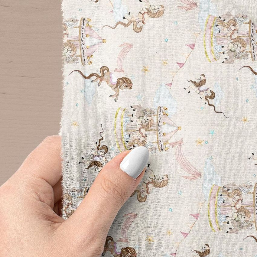 Hand holding a muslin swaddle blanket mockup with a carousel and horse pattern in soft pastel colors on a light fabric.