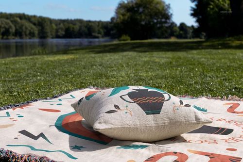 Front view of a woven pillow mockup on a decorative blanket, set on grass with a scenic lake and forest background.