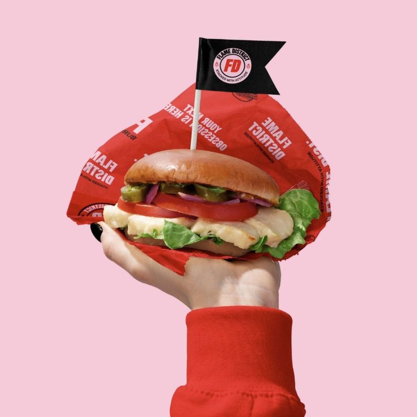 Vegan burger mockup with visible toppings and red packaging, displayed in a hand with a pink background and a logo flag.
