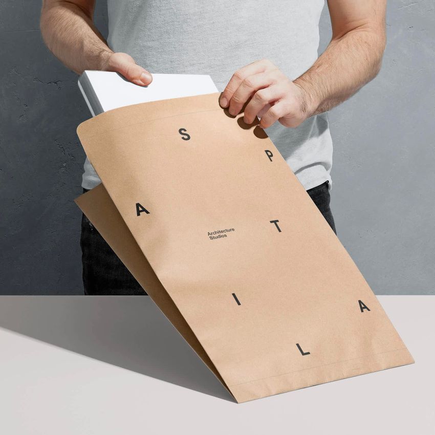 Person placing a white book into a kraft mailer mockup with bold black letters and minimal text on a gray surface.