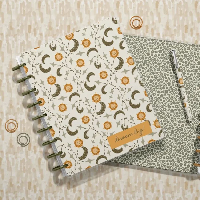 Top view of two spiral planners with floral and crescent patterns, one closed and one open, with a matching pen on a beige surface.