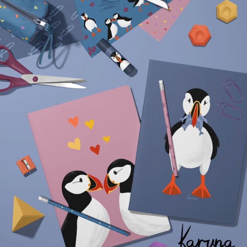 Stationery mockup featuring two classic A4 notebooks with illustrated puffins on the covers, surrounded by colorful office supplies on a blue surface.