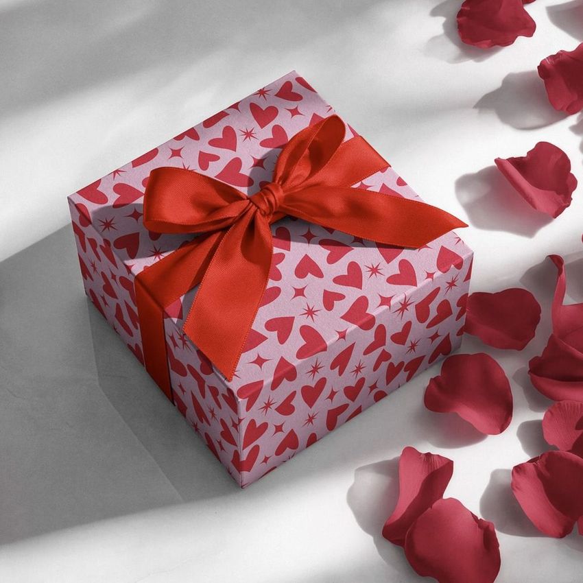 Square gift box mockup with a red ribbon, decorated with a pattern of red hearts and starbursts, surrounded by scattered rose petals.