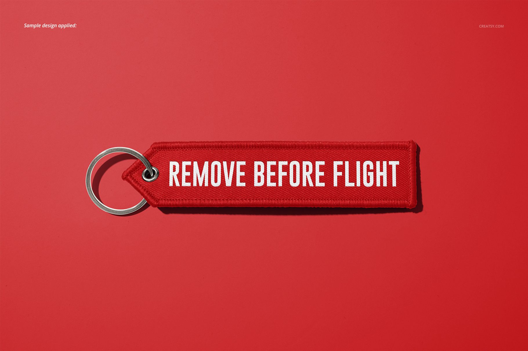 Mockup of a remove before flight keychain in red with white text, shown flat with a metal ring on a plain red surface.