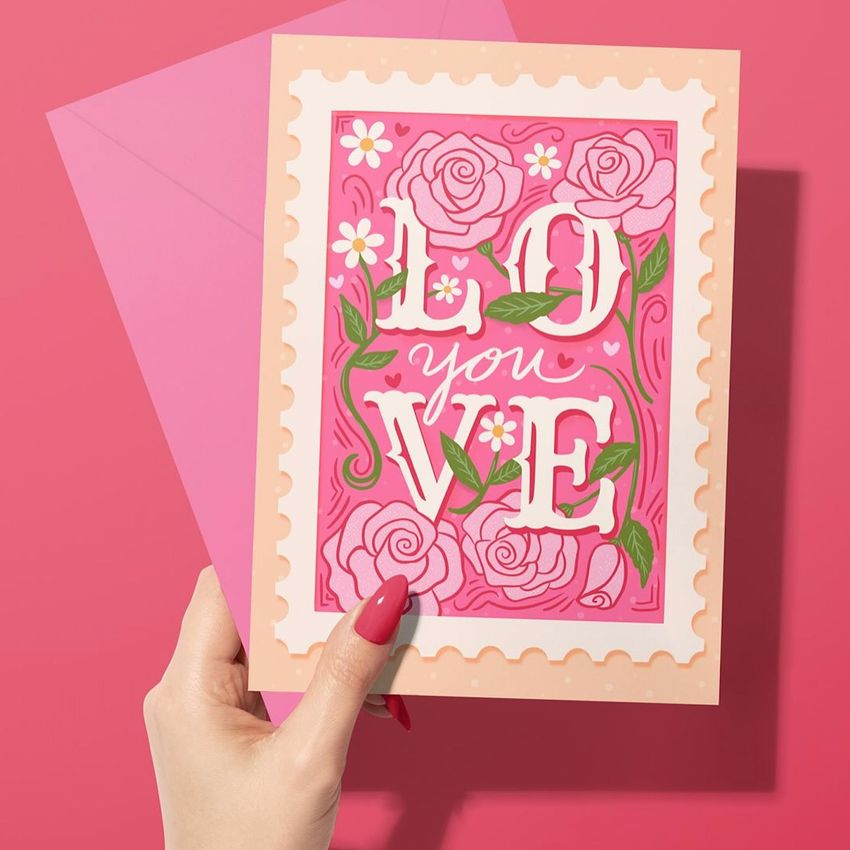 Greeting card mockup featuring a woman's hand holding a floral pink card with bold text and an envelope on a pink background.