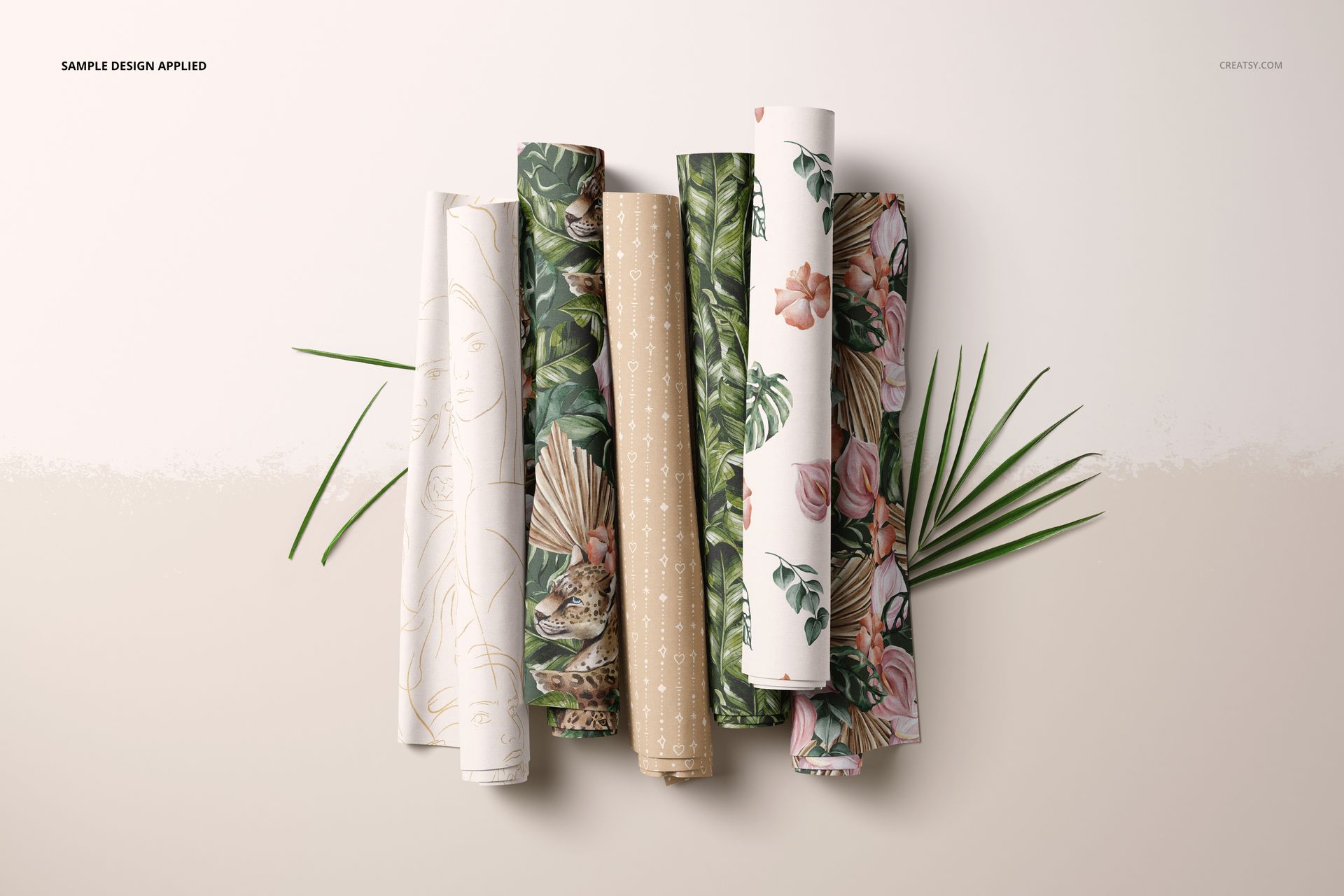 PSD mockup showing multiple fabric rolls with various nature-inspired prints, presented on a beige surface with decorative leaves.