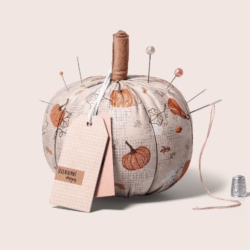 Fabric pumpkin mockup displayed as a pincushion, decorated with autumnal pumpkin and leaf patterns, pins, and sewing tags.