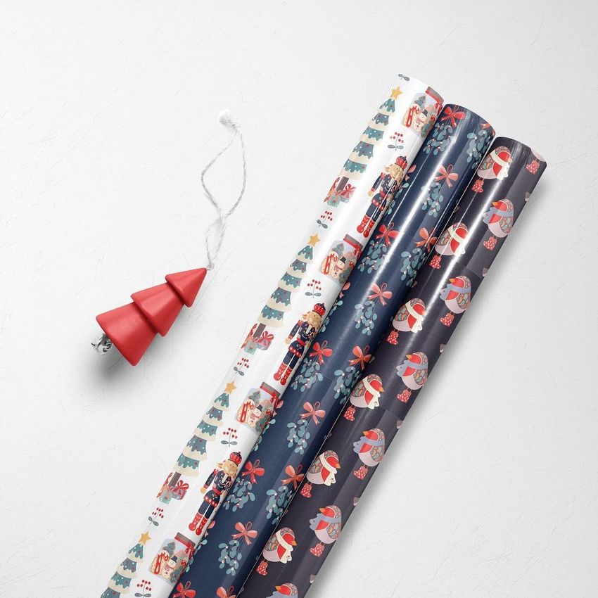 Gift wrapping paper mockup with four rolled sheets featuring festive Christmas patterns, placed on a white surface with a red tree ornament.