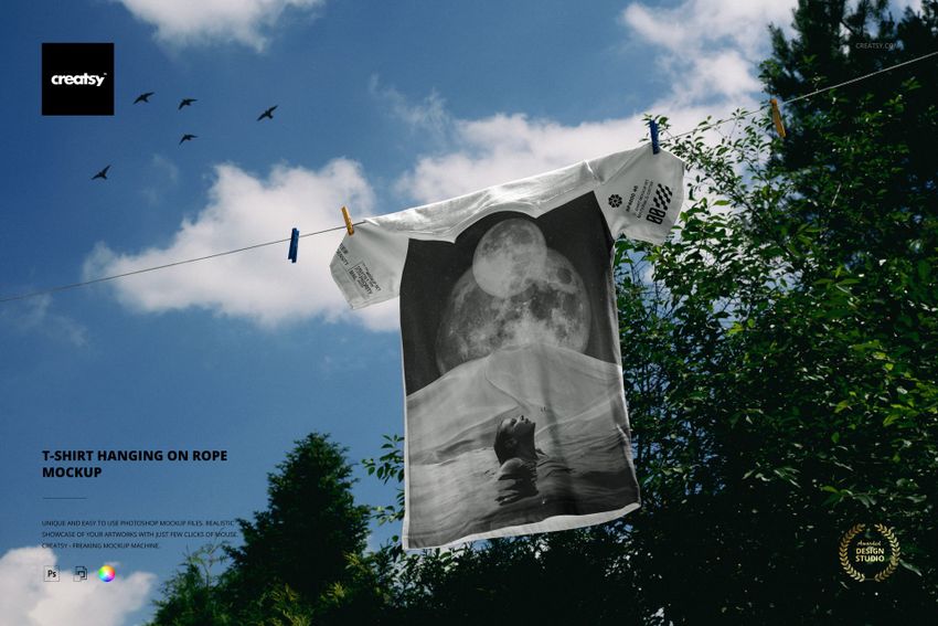 PSD mockup of a white t-shirt with a monochrome moon and mountain print, hanging on a clothesline under a partly cloudy sky.
