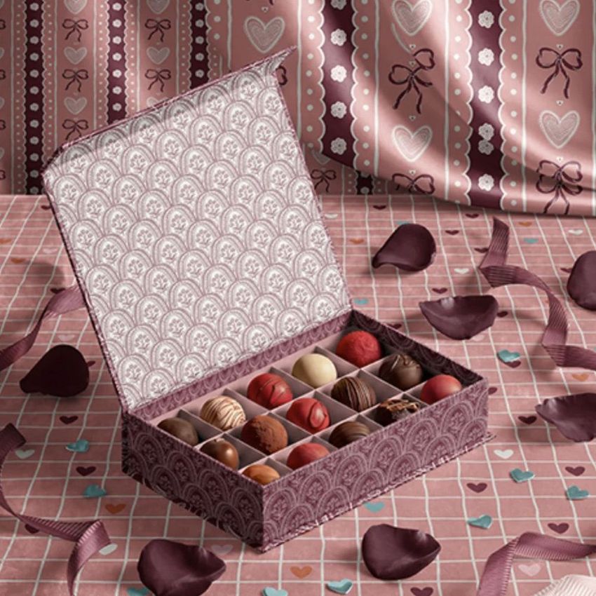 Open chocolate box mockup with assorted pralines arranged in a grid, set on a pink patterned surface with heart-shaped props.