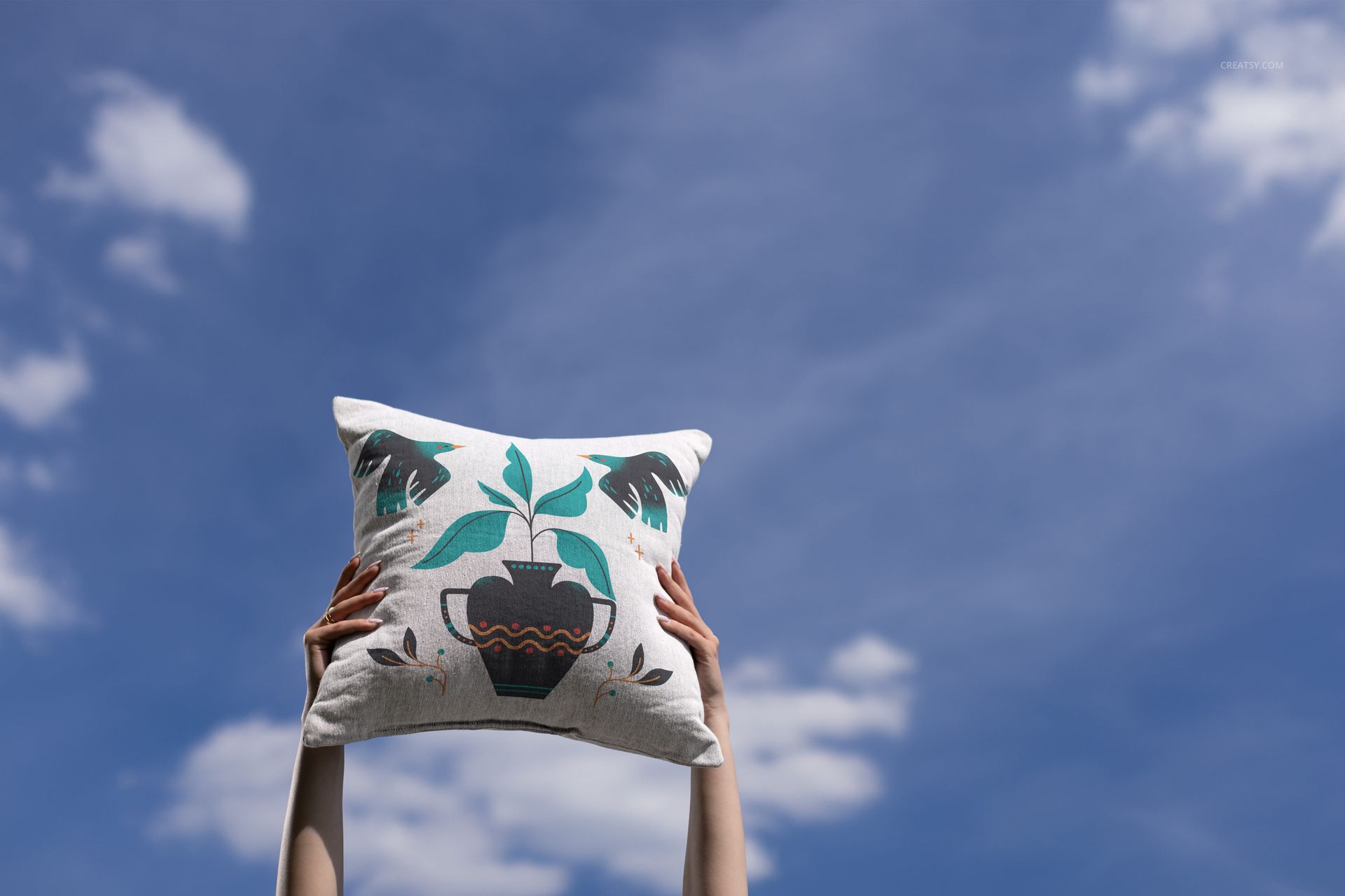 Woven pillow mockup with teal and black plant motif, raised by arms under open sky with soft white clouds.