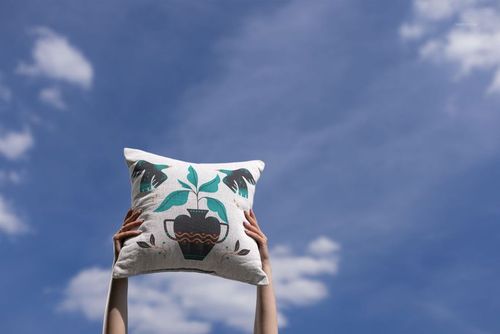Woven pillow mockup with teal and black plant motif, raised by arms under open sky with soft white clouds.