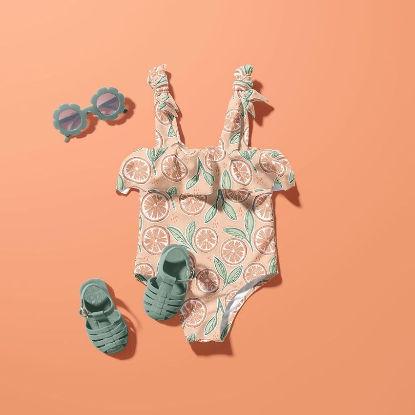 Baby swimsuit mockup with orange slice pattern displayed flat on a peach background, paired with green sandals and flower-shaped sunglasses.