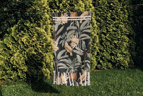 PSD mockup of a beach towel featuring a botanical design, displayed in a garden setting with tall bushes.