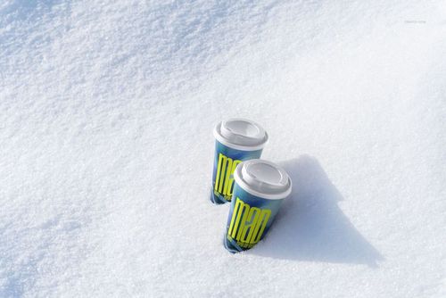 Winter-themed PSD mockup showing two drinkware paper cups with white lids and bold blue and yellow patterns in snow.