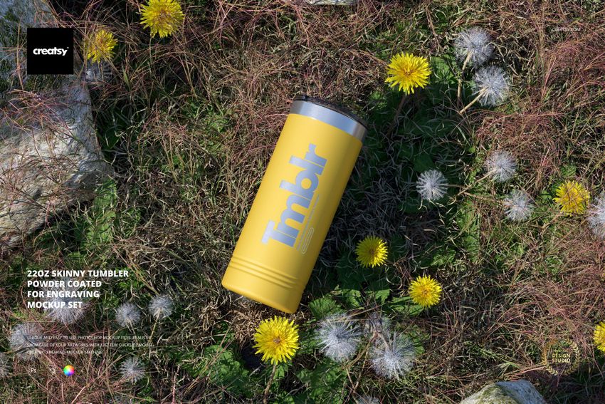 Yellow powder coated 22oz skinny tumbler lying on grass among rocks and dandelions, outdoor mockup scene.