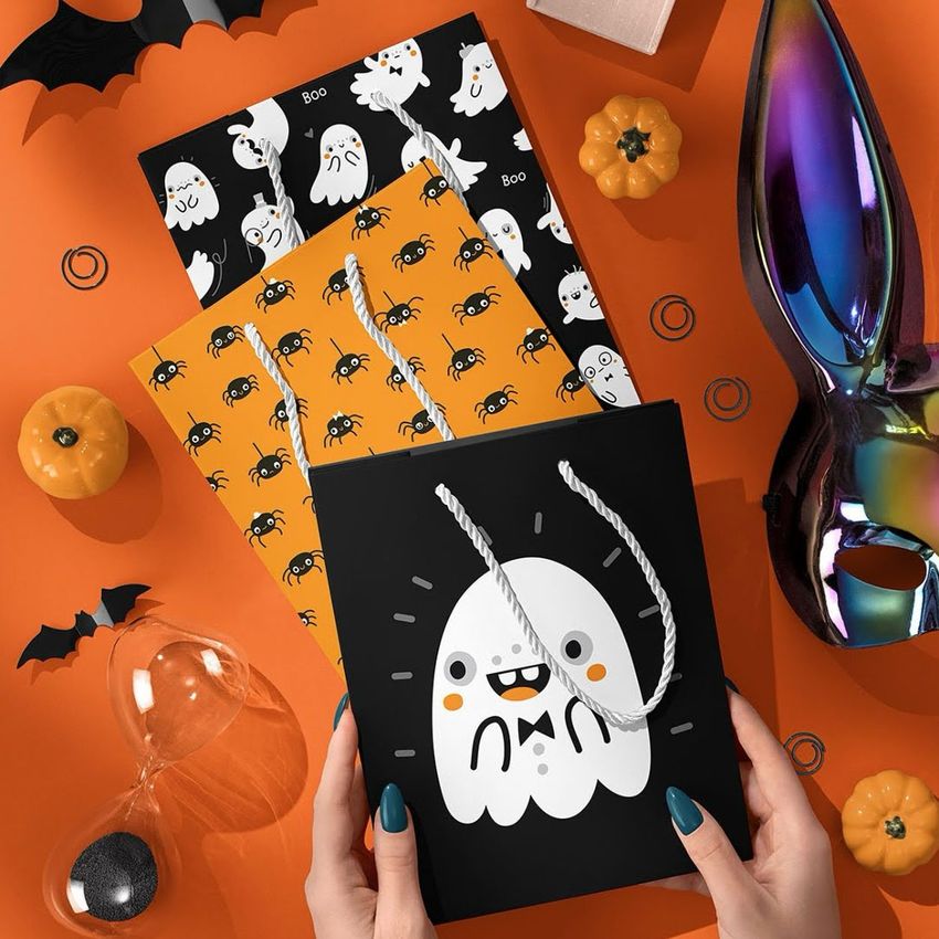 Halloween paper gift bag mockup with a cartoon ghost design, held by hands on an orange surface with themed props and patterned sheets.
