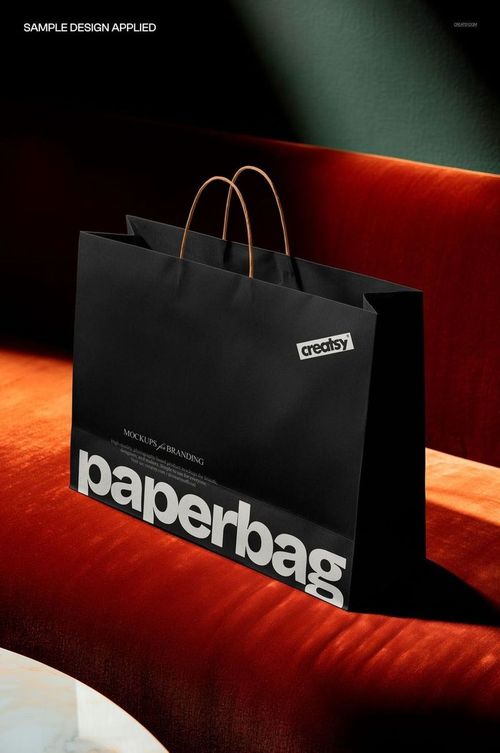 PSD mockup of a black paper shopping bag with bold white text, displayed on a deep red velvet sofa.