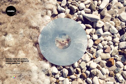 Vinyl record mockup set on rounded beach pebbles with gentle surf washing onto the shore in natural sunlight.