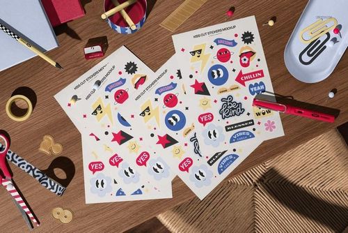 Kiss cut sticker mockup sheets with playful graphics and phrases, shown from a top view on a workspace with office supplies.