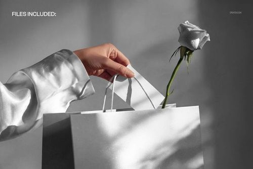 Front view of a hand holding a card above a white gift bag and rose, suitable for packaging PSD mockup presentations.